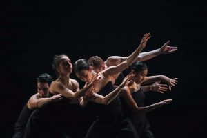 Dancers grouped together on a black stage, reaching, and looking down.