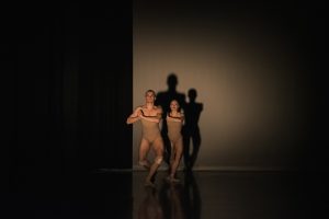 Two dancers in the spotlight on stage, in beige bodysuits, their shadows cast on the white background.