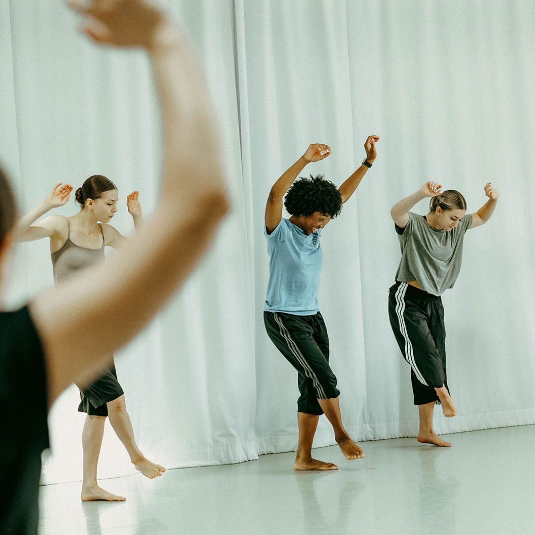 Three dancers rehearse in a bright studio with arms stretched upwards.