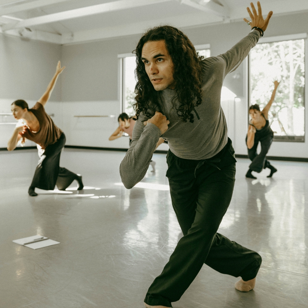 A choreographer leads a dance rehearsal in a bright studio space.
