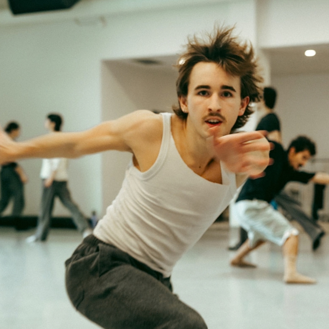 A dancer rehearses in a bright studio with an arm stretched toward the camera.